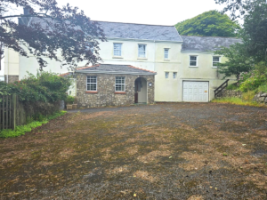Property for sale by Auction Image of a two-story house with a gray shingled roof, white exterior walls, and an attached garage. The front yard is covered with sparse greenery and a gravel driveway, and trees surround the property. The house has a stone facade section around the front door. Presented by Palace Auctions