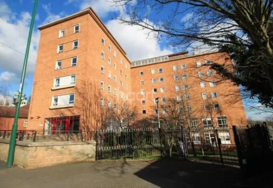Property for sale by Auction A large red-brick, multi-story building with numerous windows, surrounded by a metal fence and trees. The pathway leads to the main entrance on a sunny day with a blue sky and scattered clouds. A Day at Palace Auctions: Successful Viewings and Insightful Discussions is enhanced by the street lamp’s glow amidst the greenery. Presented by Palace Auctions