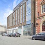 Property for sale by Auction Street view of modern building with brick and glass facade, adjacent to an older, ornate brick building. Parked cars line the street. Retail stores featuring "New Look" and "Savills" are visible on the ground floor of the modern building. Clear blue sky above. Nottingham apartment for sale by auction nearby. Presented by Palace Auctions