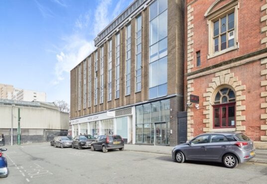 Property for sale by Auction Street view of modern building with brick and glass facade, adjacent to an older, ornate brick building. Parked cars line the street. Retail stores featuring "New Look" and "Savills" are visible on the ground floor of the modern building. Clear blue sky above. Nottingham apartment for sale by auction nearby. Presented by Palace Auctions