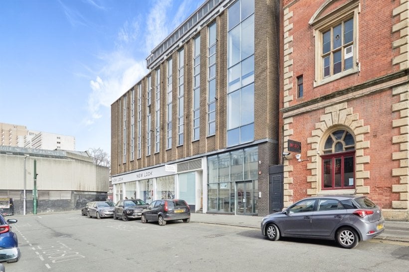 Property for sale by Auction Street view of modern building with brick and glass facade, adjacent to an older, ornate brick building. Parked cars line the street. Retail stores featuring "New Look" and "Savills" are visible on the ground floor of the modern building. Clear blue sky above. Nottingham apartment for sale by auction nearby. Presented by Palace Auctions