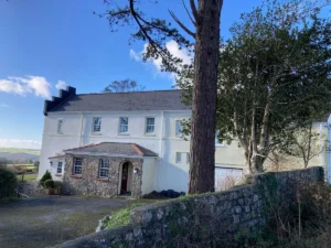 Property for sale by Auction A large, white two-story house with a stone section at the entrance, surrounded by a stone wall and trees. The sky is clear and blue, and the house is situated in a scenic, rural landscape. Presented by Palace Auctions