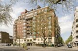 Property for sale by Auction A large, multi-story red brick apartment building stands on a corner lot, with a tree-lined street running alongside it. The building features balconies, numerous windows, and various entrances on the ground floor. On the right, another white residential building is visible. Presented by Palace Auctions