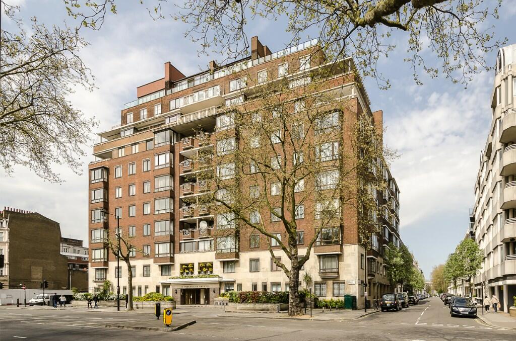 Property for sale by Auction A large, multi-story red brick apartment building stands on a corner lot, with a tree-lined street running alongside it. The building features balconies, numerous windows, and various entrances on the ground floor. On the right, another white residential building is visible. Presented by Palace Auctions