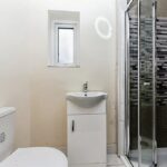 Property for sale by Auction A small bathroom featuring a white toilet, a compact vanity with a sink and faucet, a rectangular mirror above the sink, and a corner shower with a glass door. The shower has a patterned tile design in shades of grey. A small frosted window is above the sink. Presented by Palace Auctions