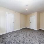 Property for sale by Auction A bright, empty room with beige walls and a grey carpet. It features two white doors and a single ceiling light. A sizable window on the left allows natural light to flood the room, highlighting the clean, minimalist design. Presented by Palace Auctions