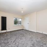 Property for sale by Auction A spacious, unfurnished room with cream-colored walls and gray carpet. The room features a small rectangular window, a black vertical radiator on the left wall, and a white door on the right wall. A single light bulb hangs from the ceiling. Presented by Palace Auctions
