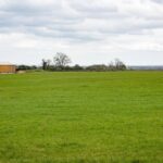 Property for sale by Auction A vast green field under a cloudy sky with a wooden and metal building on the left side. The landscape is mostly flat, with a few scattered leafless trees in the background and rolling hills visible in the distance. Presented by Palace Auctions