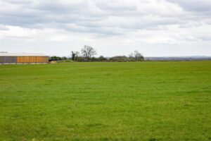 Property for sale by Auction A vast green field under a cloudy sky with a wooden and metal building on the left side. The landscape is mostly flat, with a few scattered leafless trees in the background and rolling hills visible in the distance. Presented by Palace Auctions