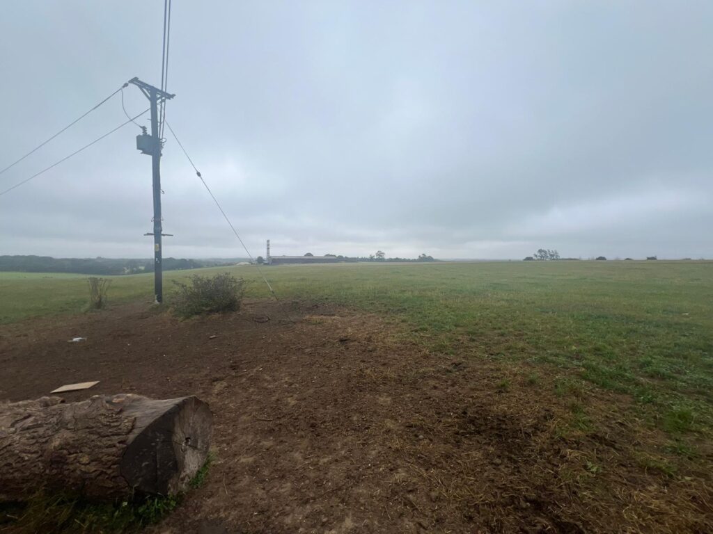 Property for sale by Auction A cloudy, overcast day in a wide, open field. The foreground features a cut tree trunk and a wooden utility pole with wires extending from it. The 2.1 acre land auction includes this prime Warlingham plot with access, where distant trees and structures shrouded in mist lie under a gray sky. Soil and grass cover the ground. Presented by Palace Auctions