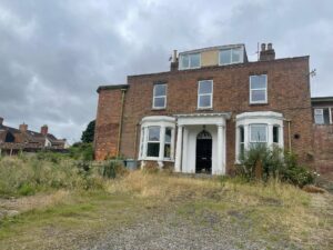 Property for sale by Auction A dilapidated two-story brick building with a prominent central entrance, white columns, and boarded windows. Overgrown grass and weeds surround the property. The sky is overcast, and neighboring houses are visible in the background. Presented by Palace Auctions