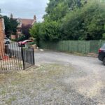 Property for sale by Auction A residential driveway with a gravel surface leads to a road. On the left is a red brick house with a black metal gate. The driveway is bordered by a green wooden fence on the right, and a few cars are parked near the road and house. Trees and greenery line the background. Presented by Palace Auctions