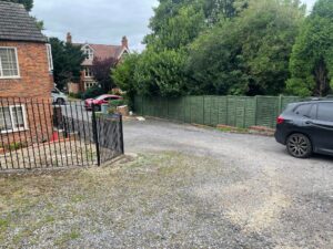 Property for sale by Auction A residential driveway with a gravel surface leads to a road. On the left is a red brick house with a black metal gate. The driveway is bordered by a green wooden fence on the right, and a few cars are parked near the road and house. Trees and greenery line the background. Presented by Palace Auctions