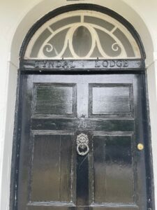 Property for sale by Auction A black wooden door with an ornate circular knocker at the center and the number 79 above it. An arched window with white trim is above the door, and "Tyndal Lodge" is inscribed above the door in the wooden frame. Presented by Palace Auctions
