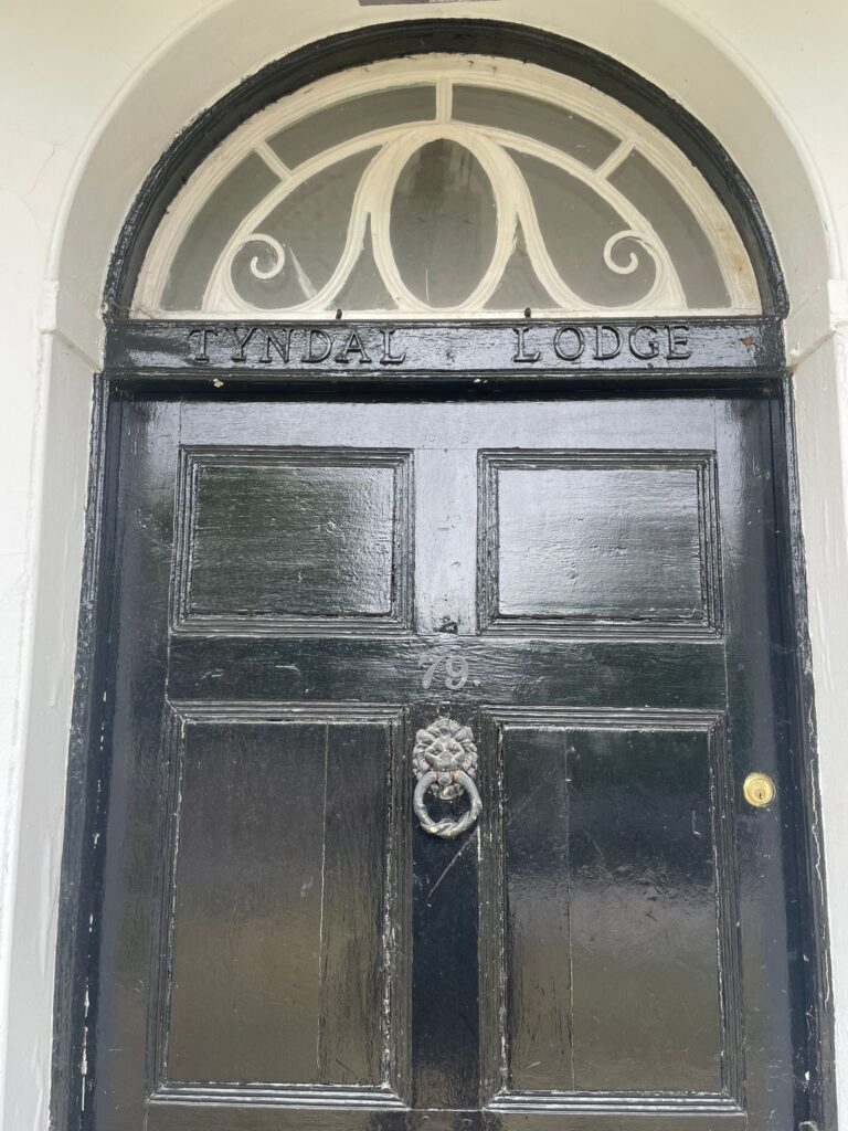 Property for sale by Auction A black wooden door with an ornate circular knocker at the center and the number 79 above it. An arched window with white trim is above the door, and "Tyndal Lodge" is inscribed above the door in the wooden frame. Presented by Palace Auctions