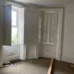 Property for sale by Auction A room with two large, curved bay windows, partially covered by interior shutters, allowing light to filter through. The room has beige carpeting and some scattered debris, including wooden planks and small items on the floor. The walls are painted white. Presented by Palace Auctions