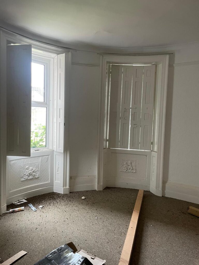 Property for sale by Auction A room with two large, curved bay windows, partially covered by interior shutters, allowing light to filter through. The room has beige carpeting and some scattered debris, including wooden planks and small items on the floor. The walls are painted white. Presented by Palace Auctions