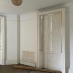 Property for sale by Auction An empty room with white walls and a high ceiling featuring a decorative light fixture. There are two tall, narrow windows and a white, panelled door. The floor is carpeted, and a blue hose and a cardboard tube are lying on the floor. Presented by Palace Auctions