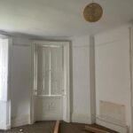 Property for sale by Auction A room with white walls and a curved corner featuring three tall windows with shutters. The floor is bare with pieces of wood and some blue tubing scattered around. A single pendant light hangs from the ceiling. Presented by Palace Auctions
