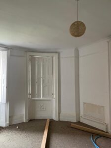 Property for sale by Auction A room with white walls and a curved corner featuring three tall windows with shutters. The floor is bare with pieces of wood and some blue tubing scattered around. A single pendant light hangs from the ceiling. Presented by Palace Auctions
