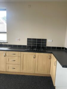 Property for sale by Auction A kitchen with light wood cabinets and dark countertops, featuring a four-burner gas stove with black tile backsplash. There are two power outlets above the countertop and a window to the left letting in natural light. The floor is dark-colored. Presented by Palace Auctions