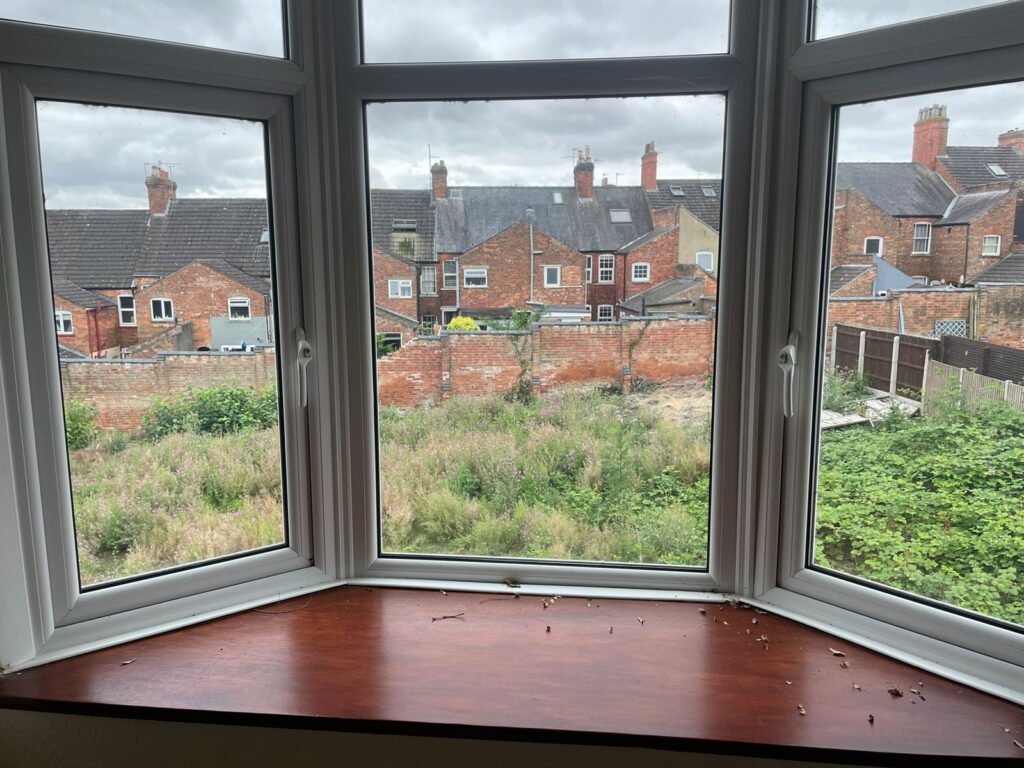 Property for sale by Auction A view through a bay window shows a backyard area overgrown with grass and weeds. Beyond the yard, a row of brick houses with chimneys lines the background beneath a cloudy sky. The window sill has some scattered debris. Presented by Palace Auctions