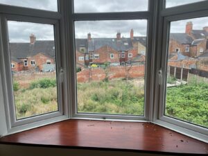 Property for sale by Auction A view through a bay window shows a backyard area overgrown with grass and weeds. Beyond the yard, a row of brick houses with chimneys lines the background beneath a cloudy sky. The window sill has some scattered debris. Presented by Palace Auctions