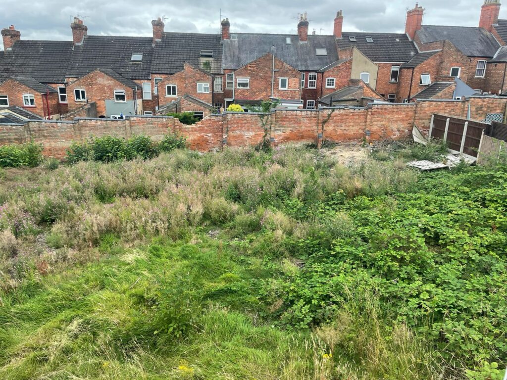 Property for sale by Auction Uneven, overgrown grassy area with patches of wildflowers and weeds. In the background, there is a row of red brick houses with chimneys. A brick wall separates the yard from the houses. The sky is overcast with grey clouds. Presented by Palace Auctions