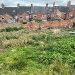 Property for sale by Auction Uneven, overgrown grassy area with patches of wildflowers and weeds. In the background, there is a row of red brick houses with chimneys. A brick wall separates the yard from the houses. The sky is overcast with grey clouds. Presented by Palace Auctions