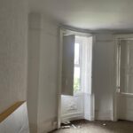 Property for sale by Auction A room with bare walls and carpeting in a state of partial renovation. Daylight streams in through two large, white-framed windows with shutters. There are scattered pieces of debris on the floor and a propped-up panel against the wall. Presented by Palace Auctions
