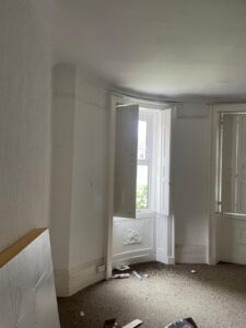 Property for sale by Auction A room with bare walls and carpeting in a state of partial renovation. Daylight streams in through two large, white-framed windows with shutters. There are scattered pieces of debris on the floor and a propped-up panel against the wall. Presented by Palace Auctions