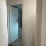 Property for sale by Auction A narrow hallway leads to an open door revealing a small, white bathroom. The bathroom has a bathtub visible in the background. The walls and door are painted white, and the hallway floor appears to be carpeted. Presented by Palace Auctions