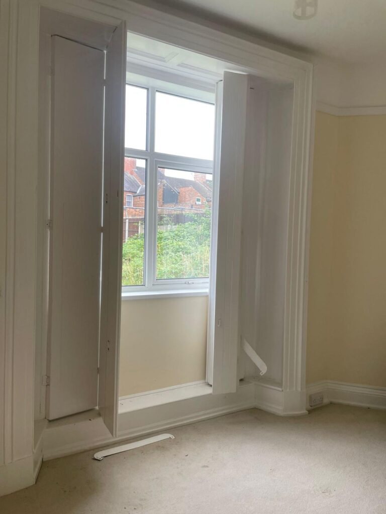 Property for sale by Auction A small room with beige walls and beige carpeting features a large window with white trim that opens up to show the view outside. The window is open and the shutters are partially open at different angles, offering a glimpse of houses and greenery in the background. Presented by Palace Auctions