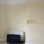 Property for sale by Auction A minimalist room with cream-colored walls and a small section of wainscoting. A blank, slightly stained wall section is visible above the wainscoting. A flat-screen TV is leaning against a radiator on the floor, with its power cable laying next to it. The carpet is light colored. Presented by Palace Auctions