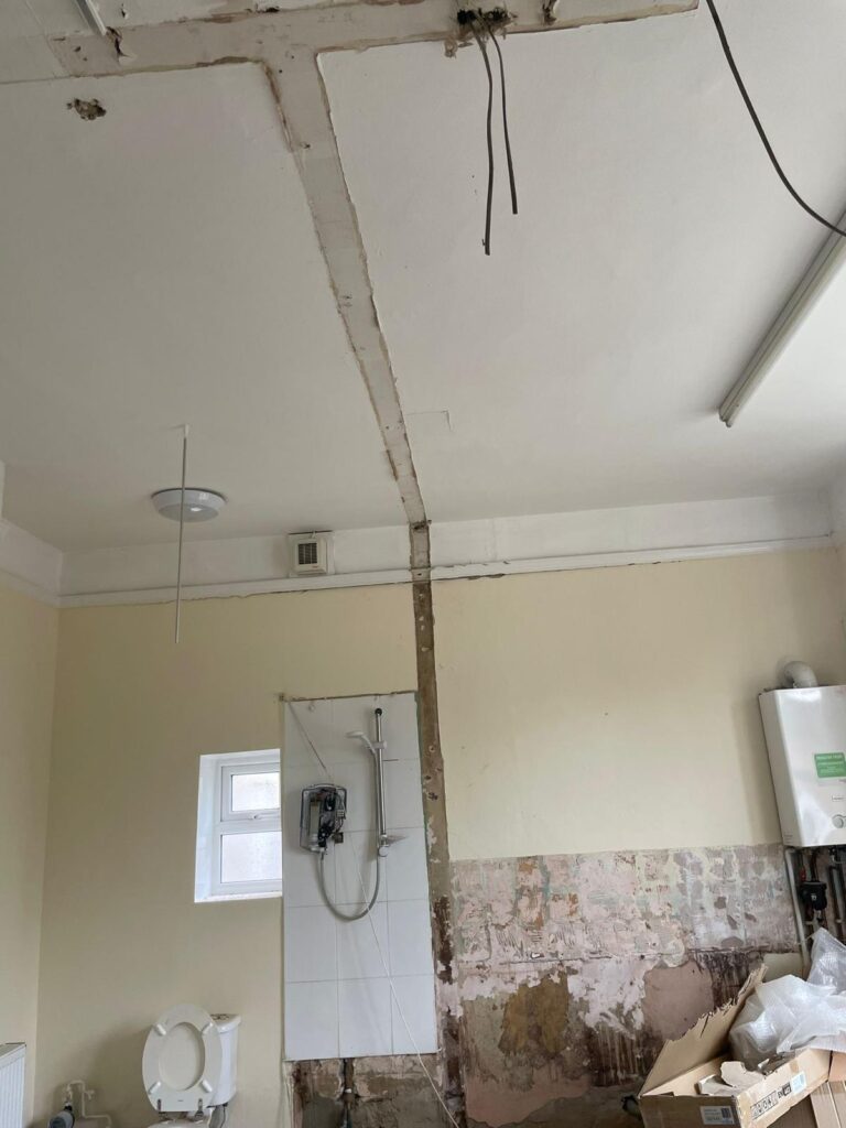 Property for sale by Auction A room under renovation, with an exposed shower unit and pipes on the wall. The ceiling has visible, unfinished seams and hanging wires. The wall has peeled paint and plaster, and construction materials are scattered on the floor. There's a toilet without a seat. Presented by Palace Auctions