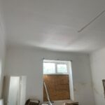 Property for sale by Auction A room with high ceilings and a boarded-up window. The walls and ceiling are off-white with some visible stains and a hole in the ceiling. Various items are scattered across the floor, including boxes, a ladder, and old furniture. Presented by Palace Auctions