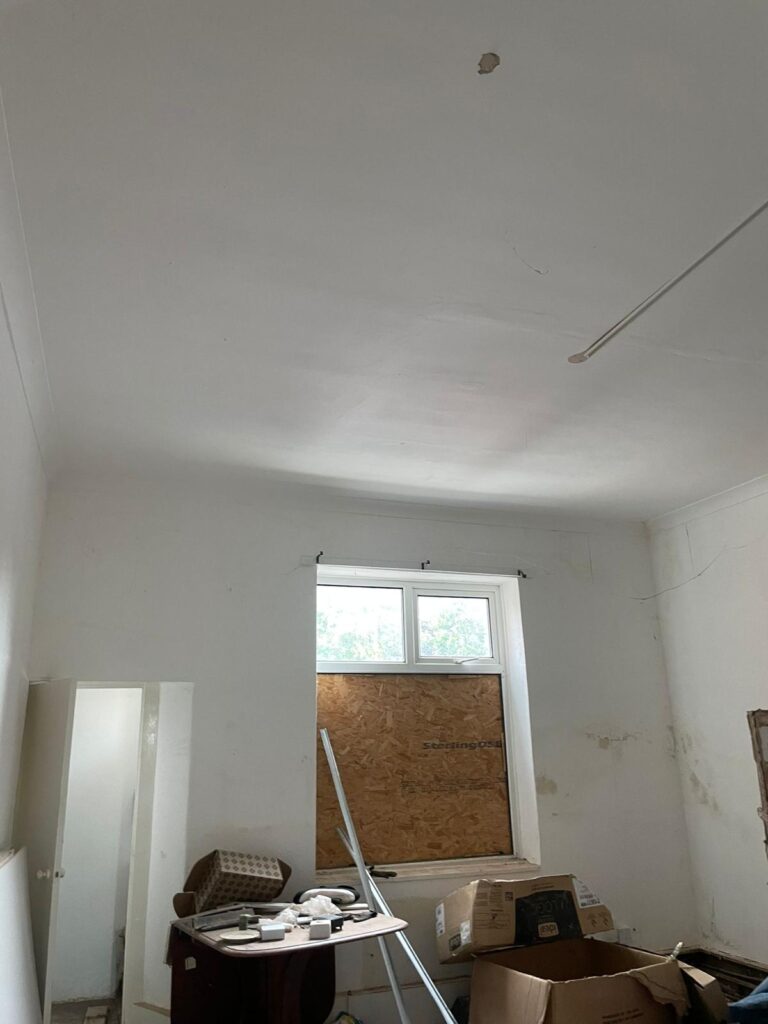 Property for sale by Auction A room with high ceilings and a boarded-up window. The walls and ceiling are off-white with some visible stains and a hole in the ceiling. Various items are scattered across the floor, including boxes, a ladder, and old furniture. Presented by Palace Auctions