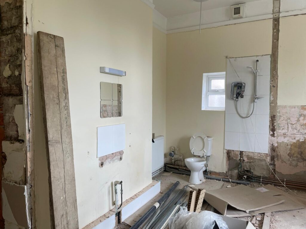 Property for sale by Auction A bathroom under renovation, with exposed pipes and walls. A toilet is installed, along with a shower area that has a partially tiled wall and an electric shower unit. Construction materials, including wooden planks and metal bars, are scattered on the floor. Presented by Palace Auctions