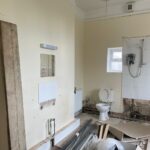 Property for sale by Auction A bathroom under renovation, with exposed pipes and walls. A toilet is installed, along with a shower area that has a partially tiled wall and an electric shower unit. Construction materials, including wooden planks and metal bars, are scattered on the floor. Presented by Palace Auctions