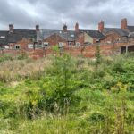 Property for sale by Auction A grassy, overgrown field with various plants and weeds in the foreground. In the background, there is a row of brick houses with multiple chimneys, some with skylights. The sky above is overcast, creating a gloomy atmosphere. Presented by Palace Auctions