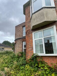 Property for sale by Auction A two-story brick building with large bay windows and white frames stands amidst overgrown vegetation. The sky is cloudy and there is another building visible in the background. The overall atmosphere is slightly abandoned and unkempt. Presented by Palace Auctions