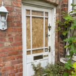 Property for sale by Auction A white wooden door with its window boarded up sits in a red brick wall. A white lantern is mounted on the left side of the wall, and the door is framed by overgrown plants. A metal letterbox and door handle are visible on the door. Presented by Palace Auctions