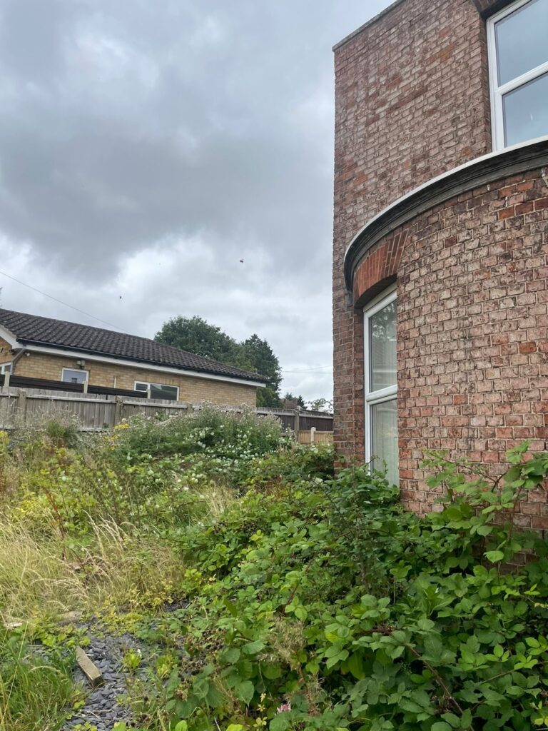 Property for sale by Auction A brick building with tall windows stands next to an overgrown garden filled with weeds and shrubs. In the background, a smaller house with a tiled roof is visible. The sky is overcast with dark clouds, indicating possible rain. Presented by Palace Auctions