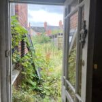 Property for sale by Auction View from an open door showing an overgrown garden with tall plants and weeds. Two ladder-like structures are leaning against a red brick wall on the left, and houses with brick chimneys are visible in the background. The sky appears overcast. Presented by Palace Auctions