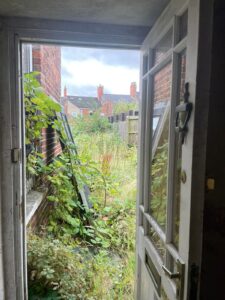 Property for sale by Auction View from an open door showing an overgrown garden with tall plants and weeds. Two ladder-like structures are leaning against a red brick wall on the left, and houses with brick chimneys are visible in the background. The sky appears overcast. Presented by Palace Auctions