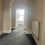 Property for sale by Auction A hallway with gray carpet leads to a bright room through an arched doorway and open double doors. The walls are beige, and a white radiator is mounted on the right-hand side. Sunlight filters into the hallway from the room ahead. Presented by Palace Auctions