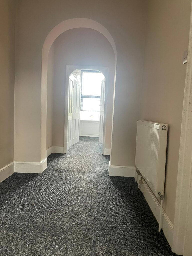 Property for sale by Auction A hallway with gray carpet leads to a bright room through an arched doorway and open double doors. The walls are beige, and a white radiator is mounted on the right-hand side. Sunlight filters into the hallway from the room ahead. Presented by Palace Auctions