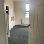 Property for sale by Auction A small, empty room with beige walls and a gray carpet. There is a single window with blinds partially drawn, allowing some natural light. A radiator is positioned below the window, and a built-in shelf unit is mounted on the left wall. The door is ajar. Presented by Palace Auctions