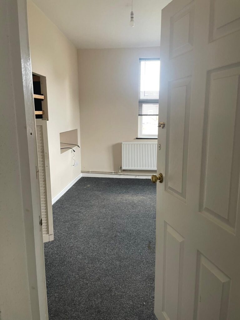 Property for sale by Auction A small, empty room with beige walls and a gray carpet. There is a single window with blinds partially drawn, allowing some natural light. A radiator is positioned below the window, and a built-in shelf unit is mounted on the left wall. The door is ajar. Presented by Palace Auctions