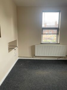 Property for sale by Auction An empty room with beige walls and a grey carpet. A window with closed blinds is on one wall above a white radiator. There are a few wall fixtures and an open cable dangling from the wall. Presented by Palace Auctions